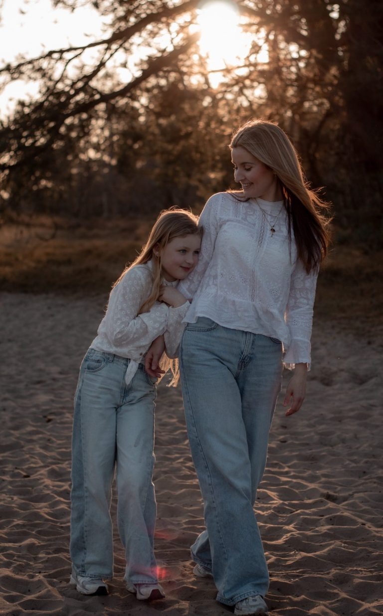 Motherhood liefdevolle shoot Twee meisjes lopen samen op het strand, omarmend met een zonsondergang op de achtergrond.
